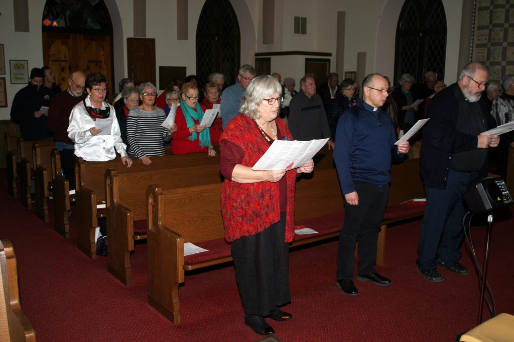 In front, from left, Rev. Dr. Christine Johnson, Fr. John Stopa and Fr. John Orban lead the community in some Christmas carols Sunday evening. Photo by Jake Davies