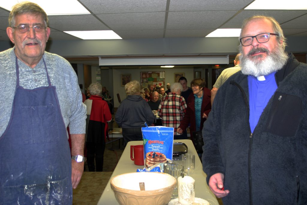 From left, volunteers Dave Rockburn and Father John Stopa mix up batter while volunteers serve pancakes in Woodlawn in the background. Photo by Jake Davies