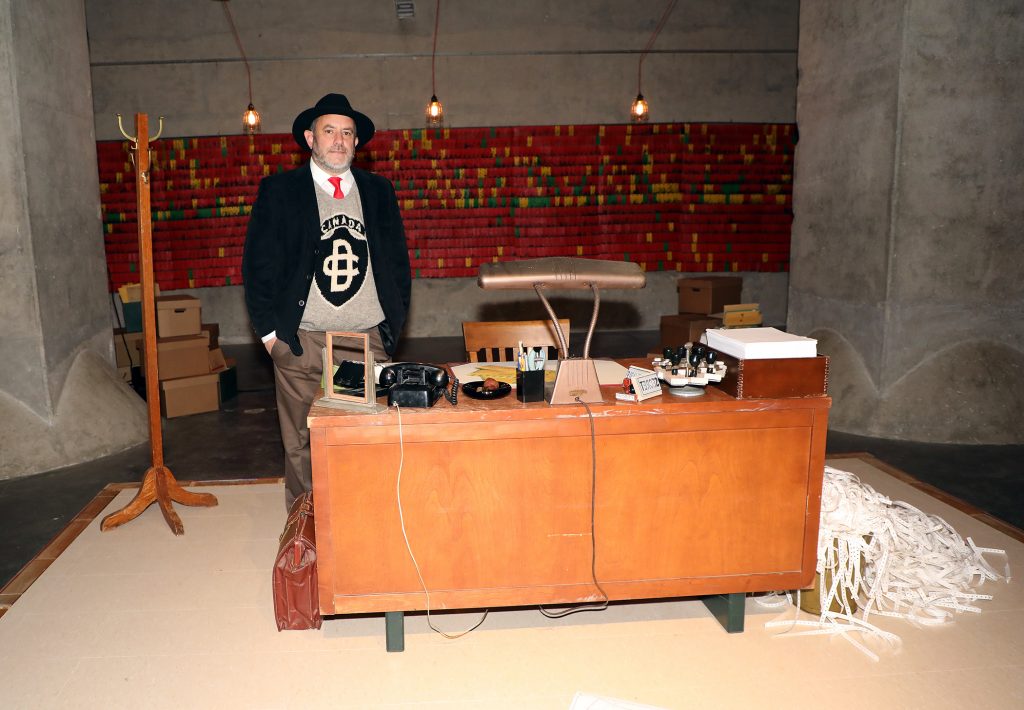 A photo of a man standing beside a desk.