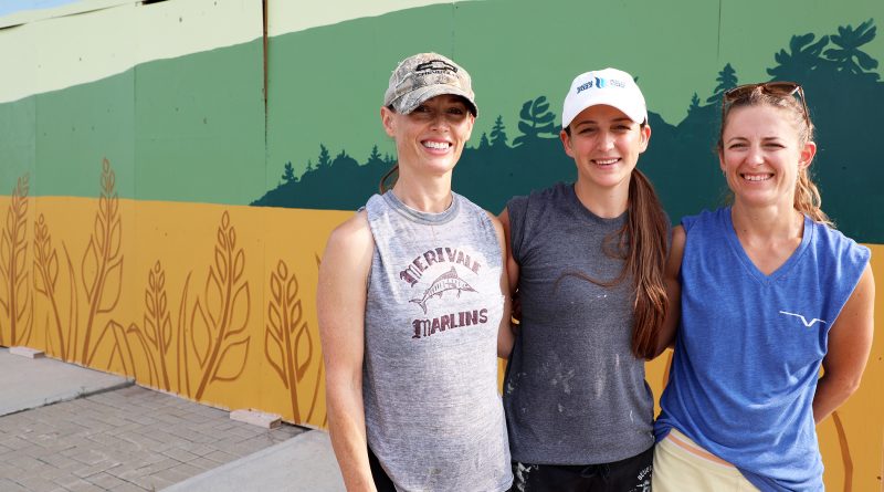 Three women pose in front of a mural.
