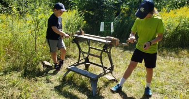 Two kids work on a sawhorse in the forest.