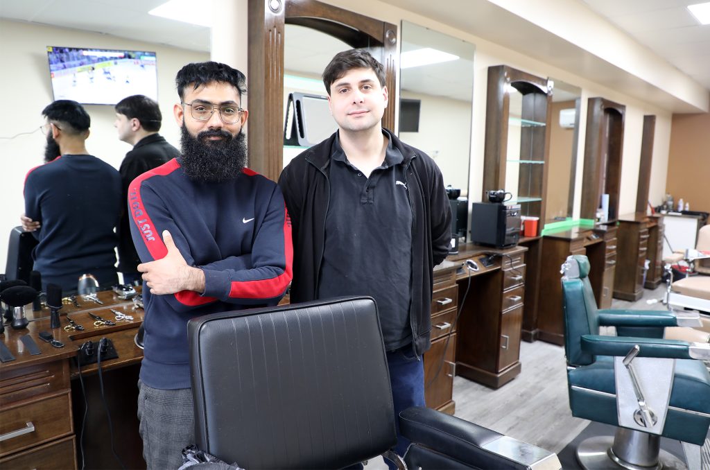 Two men pose at a barbershop chair.