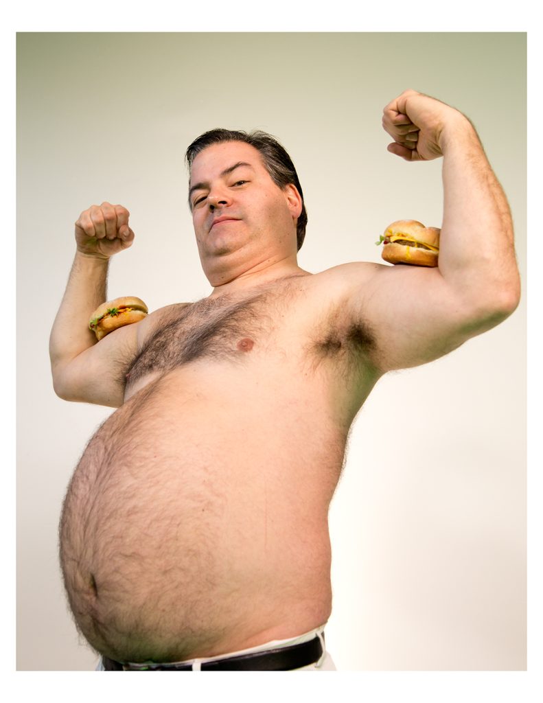 A man without a shirt flexes with two cheeseburgers on his biceps.