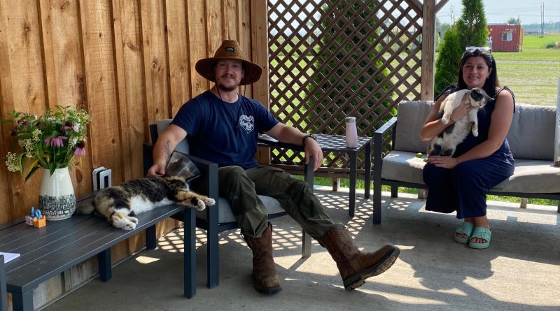 Two people pose on a porch with cats.