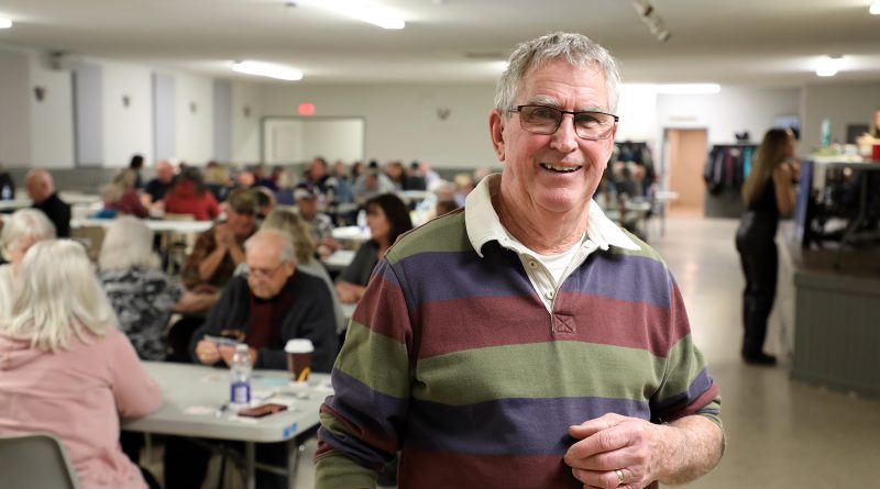 A man poses in a room full of euchre players.