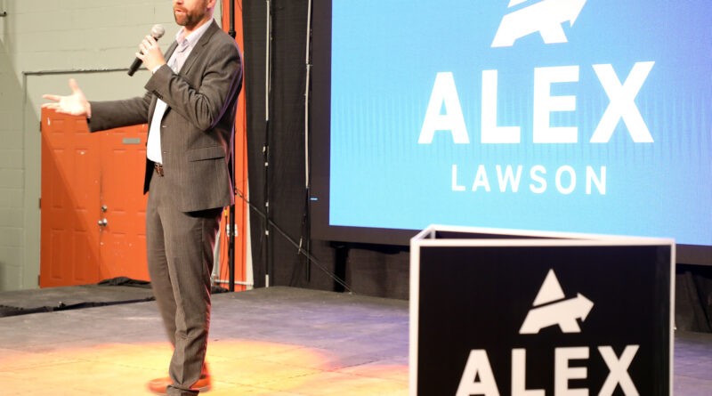 A photo of a person speaking on a stage.